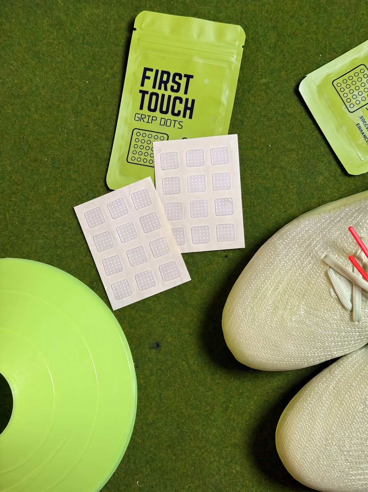 Two pairs of Grip Dots sheets and pouches on turf with a pair of soccer cleats and an agility cone